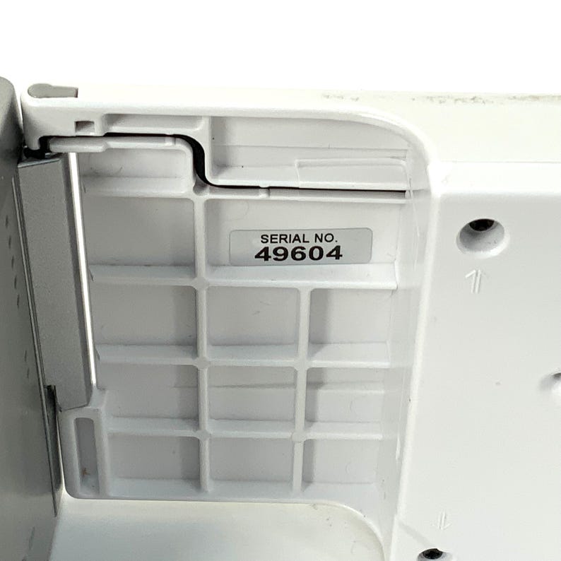 May include: Close-up of a white plastic component with a grid-like pattern. A sticker with the text "SERIAL NO. 49604" is affixed to the surface. The component has a curved edge and a metal piece.