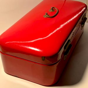 May include: A rectangular, glossy red box with a brass handle and clasps. The box has a slightly rounded top and appears to be made of metal or a similar material. The handle is a curved, ornate design.