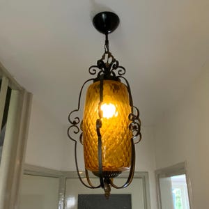 Op de afbeelding: Een vintage hanglamp van amberkleurig glas met een zwart gesmeed ijzeren frame. De lamp hangt aan een ketting en heeft een ruitvormig patroon op het glas.