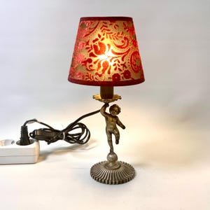 May include: A decorative table lamp with a red and gold floral lampshade. The lamp is supported by a bronze-coloured cherub figure and a circular base. The lamp includes a power cord and plug.