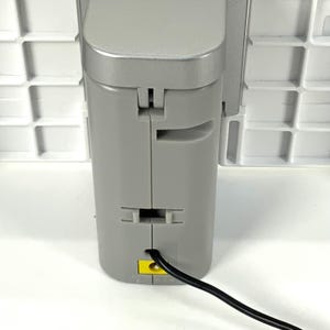 May include: A close-up of a gray electronic device with a black power cord. The device has a rectangular shape with a hinged top and a yellow port. The background is a white grid.
