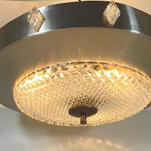 Può includere: Un lampadario da soffitto argentato con un paralume in cristallo trasparente. Il lampadario ha una base circolare con un bordo decorativo di piccoli accenti in cristallo trasparente.