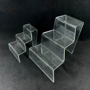 May include: Two clear acrylic display risers with three tiers each. The risers are made of clear acrylic and have a stepped design. The larger riser is on the right and the smaller riser is on the left.