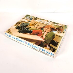 May include: A vintage Pippi Longstocking jigsaw puzzle box. The box features an image of Pippi Longstocking with her feet up on a desk. The box has the text "Pippi Langkous Jig Saw Puzzle" in blue lettering.