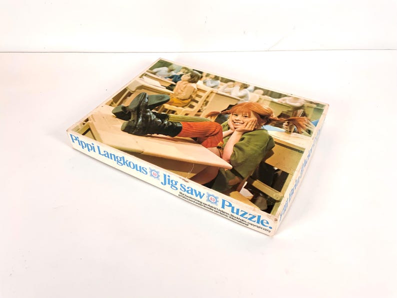 May include: A vintage Pippi Longstocking jigsaw puzzle box. The box features an image of Pippi Longstocking with her feet up on a desk. The box has the text "Pippi Langkous Jig Saw Puzzle" in blue lettering.
