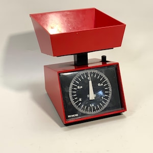 May include: A retro red kitchen scale featuring a black dial with white numerals and markings. The scale has a red tray and a maximum capacity of 1000 grams. The dial shows measurements in grams.