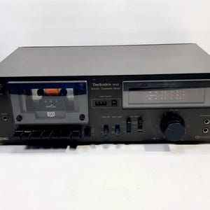 Op de afbeelding: Een zwarte Technics M12 stereo cassette deck. De deck heeft een cassettebandje zichtbaar in het tape compartiment. Het voorpaneel heeft diverse knoppen, een volumeknop en een digitale display. De tekst "Technics M12 Stereo Cassette Deck" is zichtbaar.