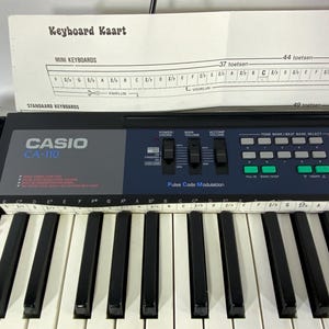 May include: A Casio CA-110 electronic keyboard with black and white keys. The keyboard has a control panel with various buttons and sliders. A sheet of paper with the text "Keyboard Kaart" is above the keyboard.