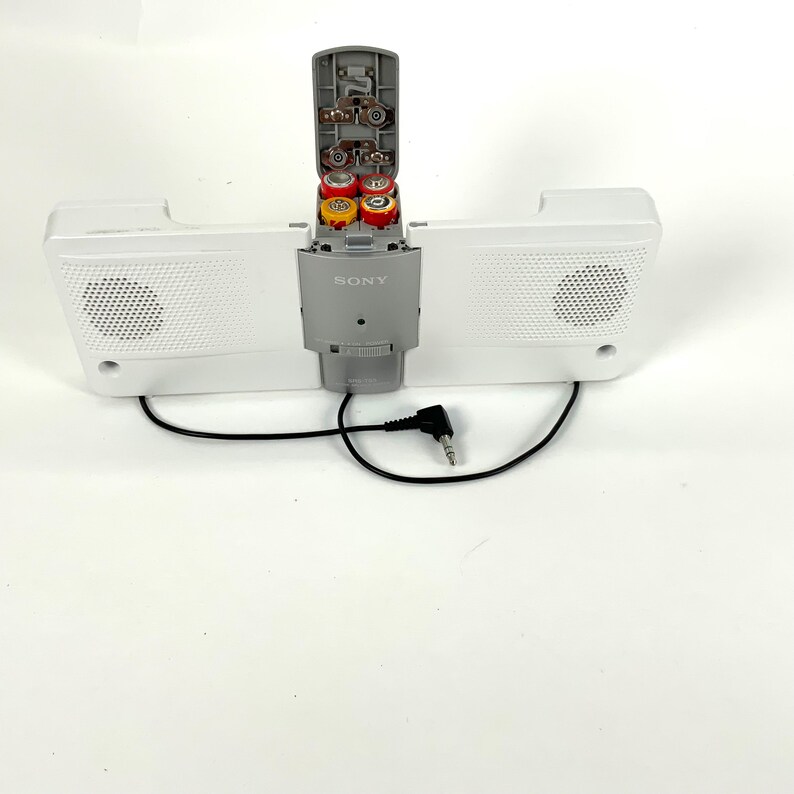 May include: A white Sony speaker system with two speakers and a central control unit. The control unit is open, revealing several batteries. A black cable with a 3.5mm jack extends from the unit.