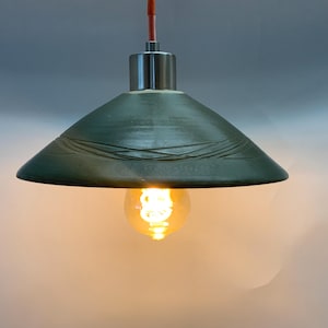 Puede incluir: Una lámpara colgante de cerámica verde con un diseño simple y moderno. La lámpara tiene una pantalla ancha y plana con una superficie texturizada y una sola bombilla.