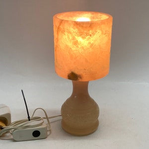 May include: A white alabaster lamp with a carved floral design. The lamp has a round base and a cylindrical shade. The lamp is lit and casting a warm glow.