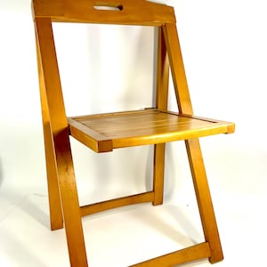 May include: A light brown wooden folding chair with a rectangular seat and a backrest featuring a handle cutout. The chair has angled legs and a simple, functional design. The chair is made of wood.