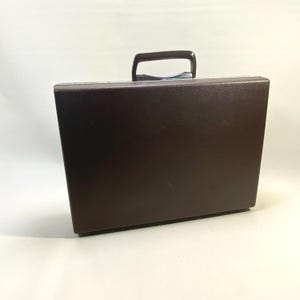 Puede incluir: Un maletín marrón con asa plateada. La funda rectangular está hecha de un material liso y tiene un diseño sencillo y clásico. El asa es curva y está sujeta a la parte superior de la funda. El maletín está sobre un fondo blanco.