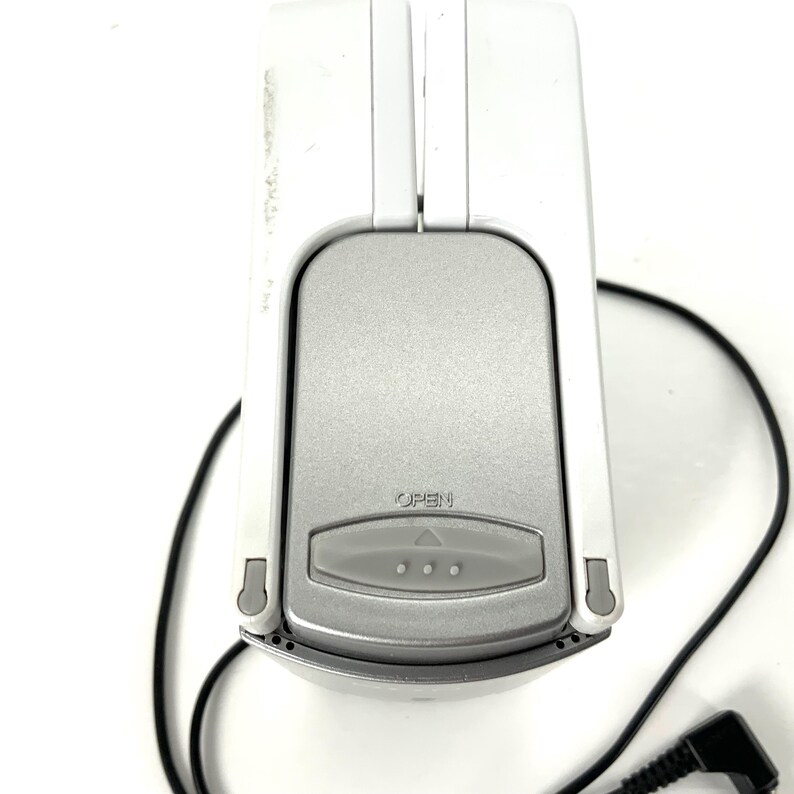 May include: A white and silver electronic device with a black cord. The device has a silver center with the word "OPEN" and three small dots. The top and sides are white. The cord has a right-angle connector.