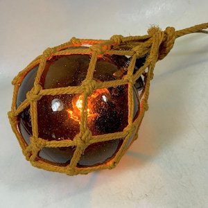 Puede incluir: Una boya de vidrio marrón con una red de cuerda, iluminada desde el interior con un cálido resplandor naranja.