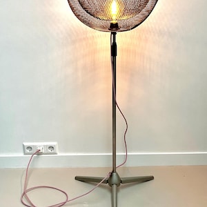 May include: A vintage-style floor lamp with a dark metal mesh shade. The lamp has a silver-colored stand with a tripod base and a pink power cord. The light bulb is visible inside the shade, casting a warm glow.
