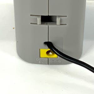 May include: Close-up of a gray electronic device with a black power cord. The device has a yellow port and the text "DC IN 6V". The device has a slot and two tabs.