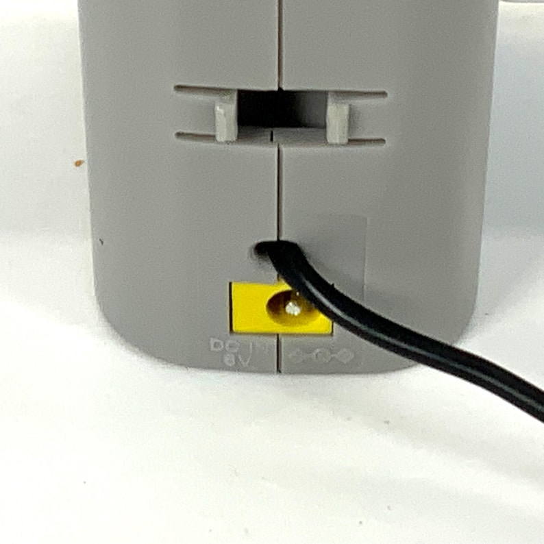 May include: Close-up of a gray electronic device with a black power cord. The device has a yellow port and the text "DC IN 6V". The device has a slot and two tabs.