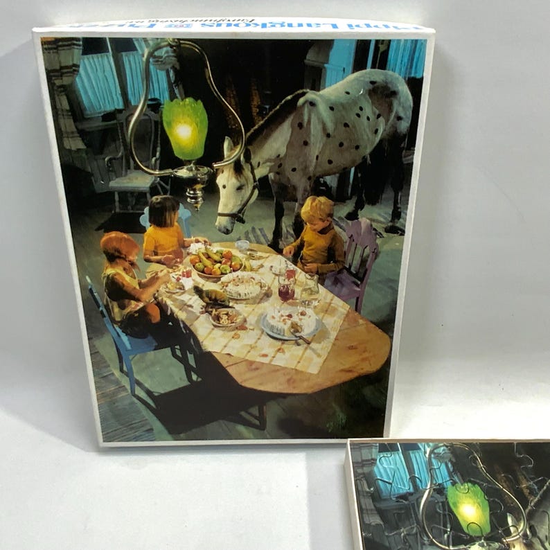 May include: A rectangular jigsaw puzzle box featuring an illustration of two children sitting at a wooden dining table with a white horse standing beside them. A green glass pendant light hangs above the table, which is set with plates of food.