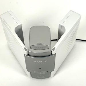 May include: A white and gray Sony battery charger. The charger has two white wings that open and close, with a gray center section. The Sony logo is visible on the front. The model number is visible on the front.