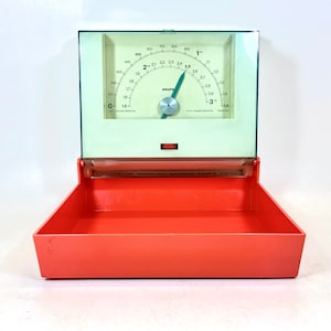 Op de afbeelding: Een vintage oranje en witte keukenweegschaal met een glazen bovenkant en een wijzerplaat die tot 3 kilogram aangeeft. De weegschaal is gemaakt door Krups.