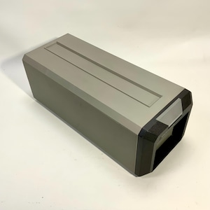 May include: A rectangular, gray speaker with a recessed top and black accents. The speaker has a modern design and is likely used for audio playback. The front corners are black, and the top has two recessed lines.