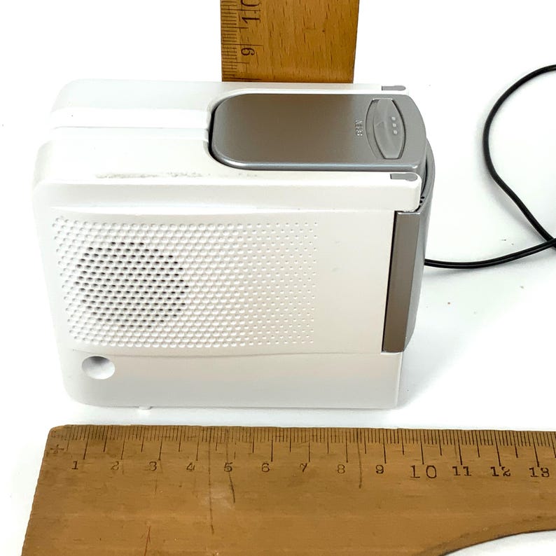 May include: A white and gray portable radio with a speaker grill, a flip-up top, and a silver-colored control panel. A wooden ruler is placed on top of the radio. The radio measures approximately 4 inches in height.
