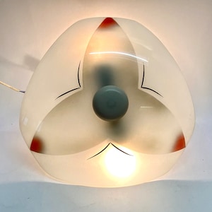 Puede incluir: Una lámpara de techo de vidrio blanco esmerilado con un diseño de tres hojas. Las hojas tienen líneas negras y acentos rojos. La lámpara tiene un centro gris con una bombilla en el interior.