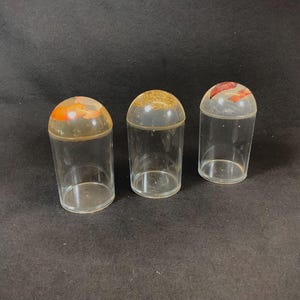 Könnte beinhalten: Drei durchsichtige Glasbehälter mit unterschiedlich farbigen Deckeln. Die Deckel sind hellorange, gold und rot.