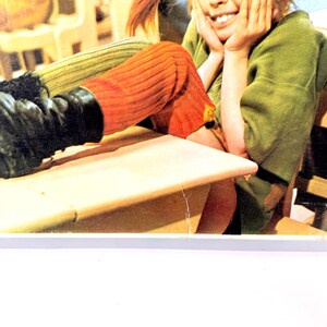 May include: A person wearing orange and green knit leg warmers and a green top. The person is smiling and resting their head on their hands. Black boots are visible.