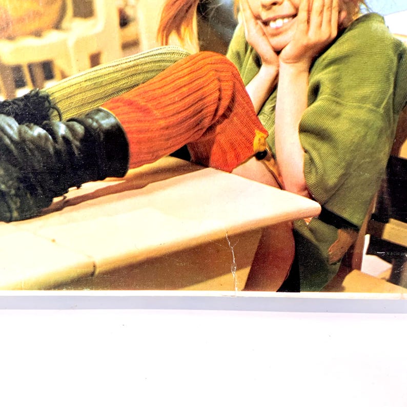 May include: A person wearing orange and green knit leg warmers and a green top. The person is smiling and resting their head on their hands. Black boots are visible.