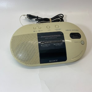 May include: A beige Sony Dream Machine clock radio with a black digital display and a speaker grille. The radio has buttons for sleep, alarm, radio, and buzzer. The text "DREAM MACHINE" is printed on the front of the radio.