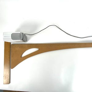 May include: A wooden drafting square and a curved ruler are positioned on a white surface. A small, white and gray device with a black cord is also present. The drafting square is approximately 12 inches long.