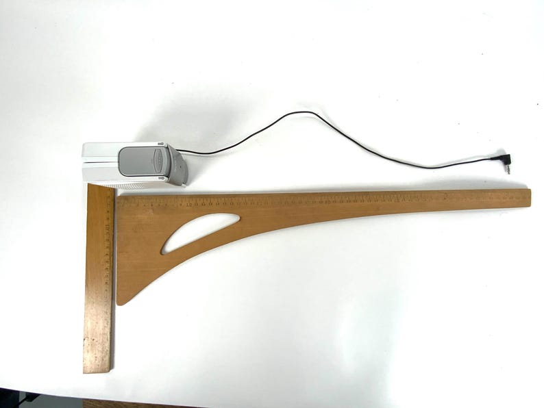 May include: A wooden drafting square and a curved ruler are positioned on a white surface. A small, white and gray device with a black cord is also present. The drafting square is approximately 12 inches long.