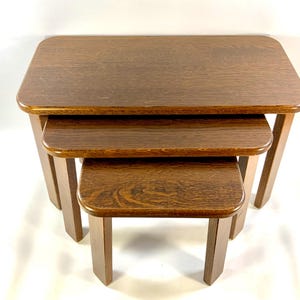 May include: Set of three nesting tables made of dark brown wood. Each table has a rectangular top with rounded corners and four straight legs. The tables are designed to fit inside each other for storage.
