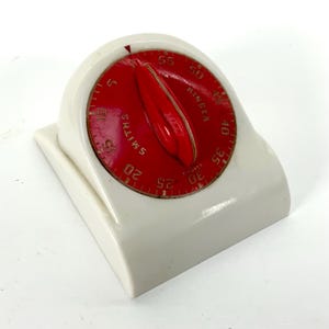 Może przedstawiać: Zegar kuchenny w stylu vintage z czerwoną tarczą i białą podstawą. Tarcza posiada czerwoną wskazówkę i złote cyfry wskazujące minuty. Zegar jest oznaczony napisem "SMITHS RINGER".