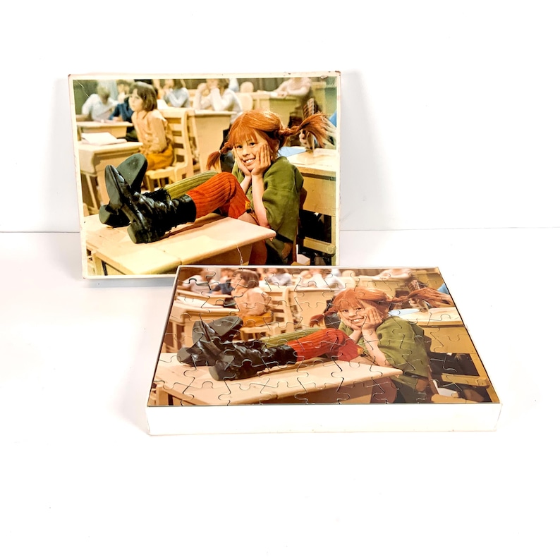 May include: Two vintage jigsaw puzzles featuring a colorful image of a young girl with red pigtails, wearing a green jacket and black boots, sitting at a school desk. The puzzles are in their original boxes.