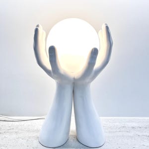 Puede incluir: Lámpara de mesa de cerámica blanca con una luz esférica brillante sostenida por dos manos estilizadas. La lámpara tiene un acabado liso y brillante y un diseño moderno. Es una lámpara decorativa.