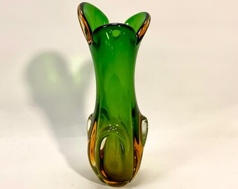 May include: A green glass vase with a distinctive form. The vase features a flared top with two upward-curving elements and a rounded base. The edges are highlighted with a golden-brown hue, enhancing the design. The vase is empty.