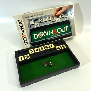 May include: A black game board with a green felt interior. The game is called "Down & Out" and the goal is to have the lowest score. The board has numbered slots and dice are used to play. The box for the game is in the background.