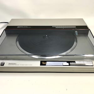 Może przedstawiać: Srebrny gramofon Technics SL-QL3 z napędem bezpośrednim i przezroczystą osłoną przeciwpyłową. Gramofon ma czarną płytę na talerzu i czarny przewód zasilający. Przedni panel ma przyciski i napis "Technics".