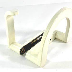 May include: White plastic toilet paper holder with a curved design. The holder is attached to a wall with screws.