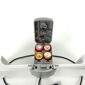 May include: A gray Sony device with an open battery compartment containing four AA batteries. The batteries are red and yellow. The device has a white frame and a black cable.