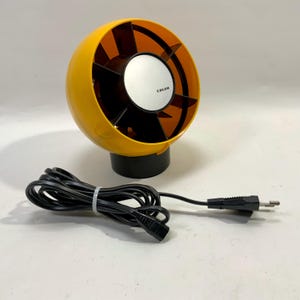 May include: A spherical desk fan with a bright yellow outer casing and a black interior housing multiple blades. The centre features a circular silver cap with the black text CALOR printed on it. A black power cord with a plug is attached to the base.