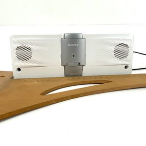 May include: A white Sony speaker dock with a silver control panel. The speaker has two circular speaker grills and a black power cord. A wooden drafting square rests in front of the speaker.