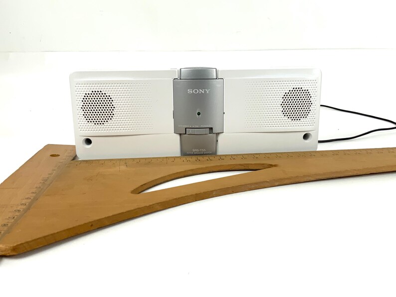 May include: A white Sony speaker dock with a silver control panel. The speaker has two circular speaker grills and a black power cord. A wooden drafting square rests in front of the speaker.