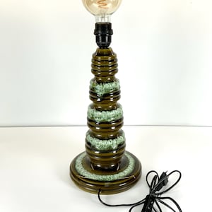 Puede incluir: Una lámpara de mesa de cerámica con acabado esmaltado marrón y verde. La lámpara tiene un diseño cilíndrico apilado con una base redonda y una bombilla grande y redonda. El cable y el enchufe de la lámpara son negros.
