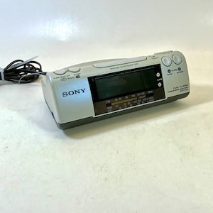 Puede incluir: Un radio despertador dual alarma Sony blanco con una pantalla digital. El radio despertador tiene un temporizador de apagado, un botón de repetición y ajustes de alarma. La radio tiene bandas AM y FM. El radio despertador está conectado a la red eléctrica con un cable de alimentación.