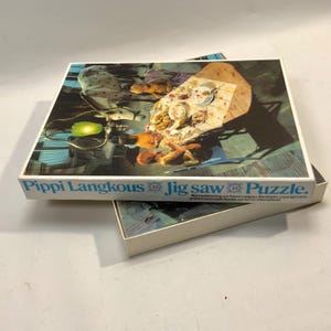 May include: A rectangular cardboard jigsaw puzzle box featuring the text Pippi Langkous Jig saw Puzzle. The box lid displays a vintage illustration of a dining scene with a horse, a table with food, and figures. The box is open, revealing puzzle pieces.