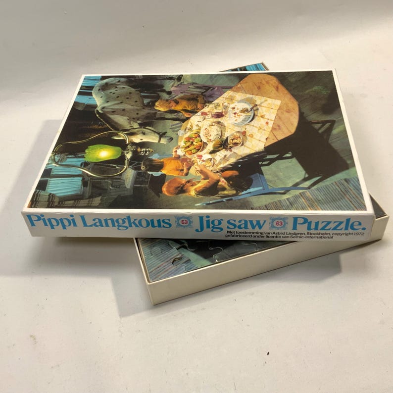 May include: A rectangular cardboard jigsaw puzzle box featuring the text Pippi Langkous Jig saw Puzzle. The box lid displays a vintage illustration of a dining scene with a horse, a table with food, and figures. The box is open, revealing puzzle pieces.